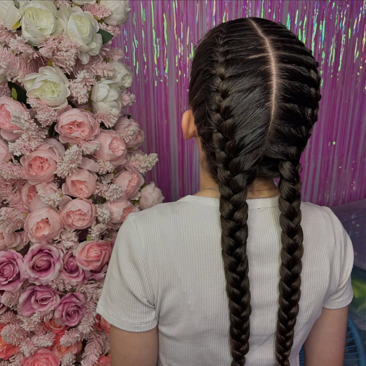 Person showing two long center-parted braids against flowers and a purple curtain