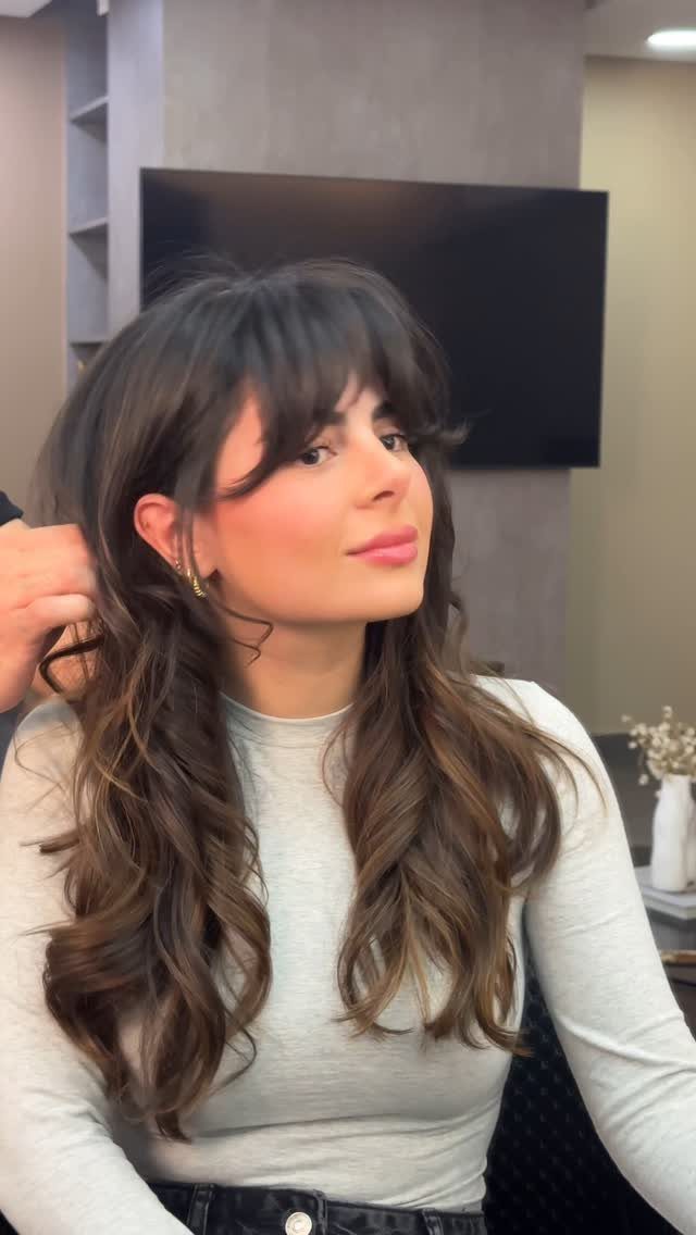 Woman with long wavy brown hair and eyebrow-grazing bangs in a salon