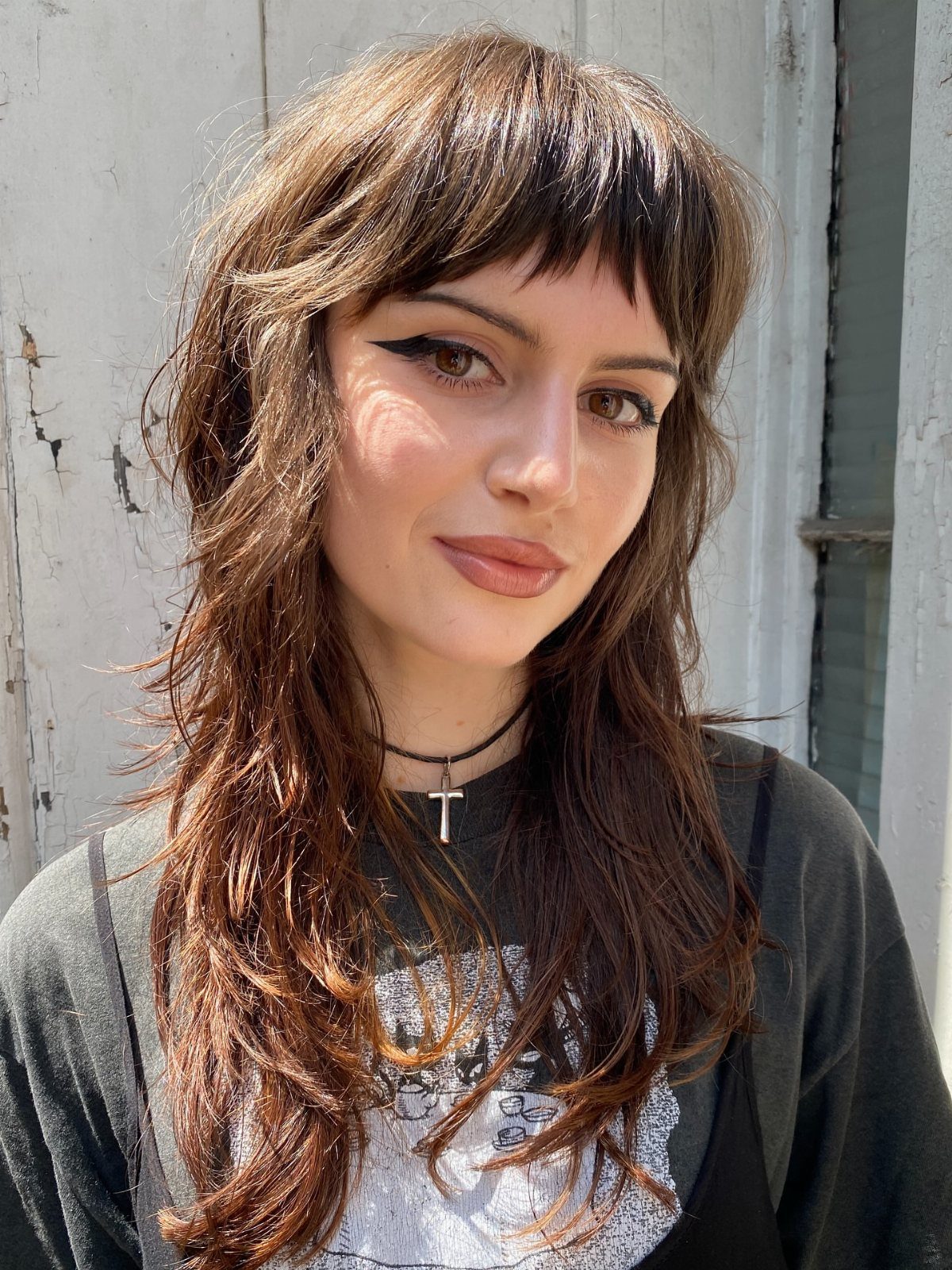 Woman with long layered brown hair and textured bangs posing against a gray wall