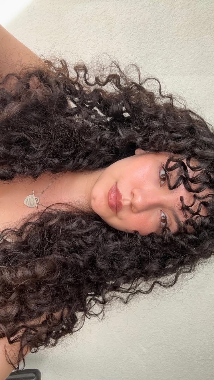 Person with long dark curls and curly bangs posing against a pale wall