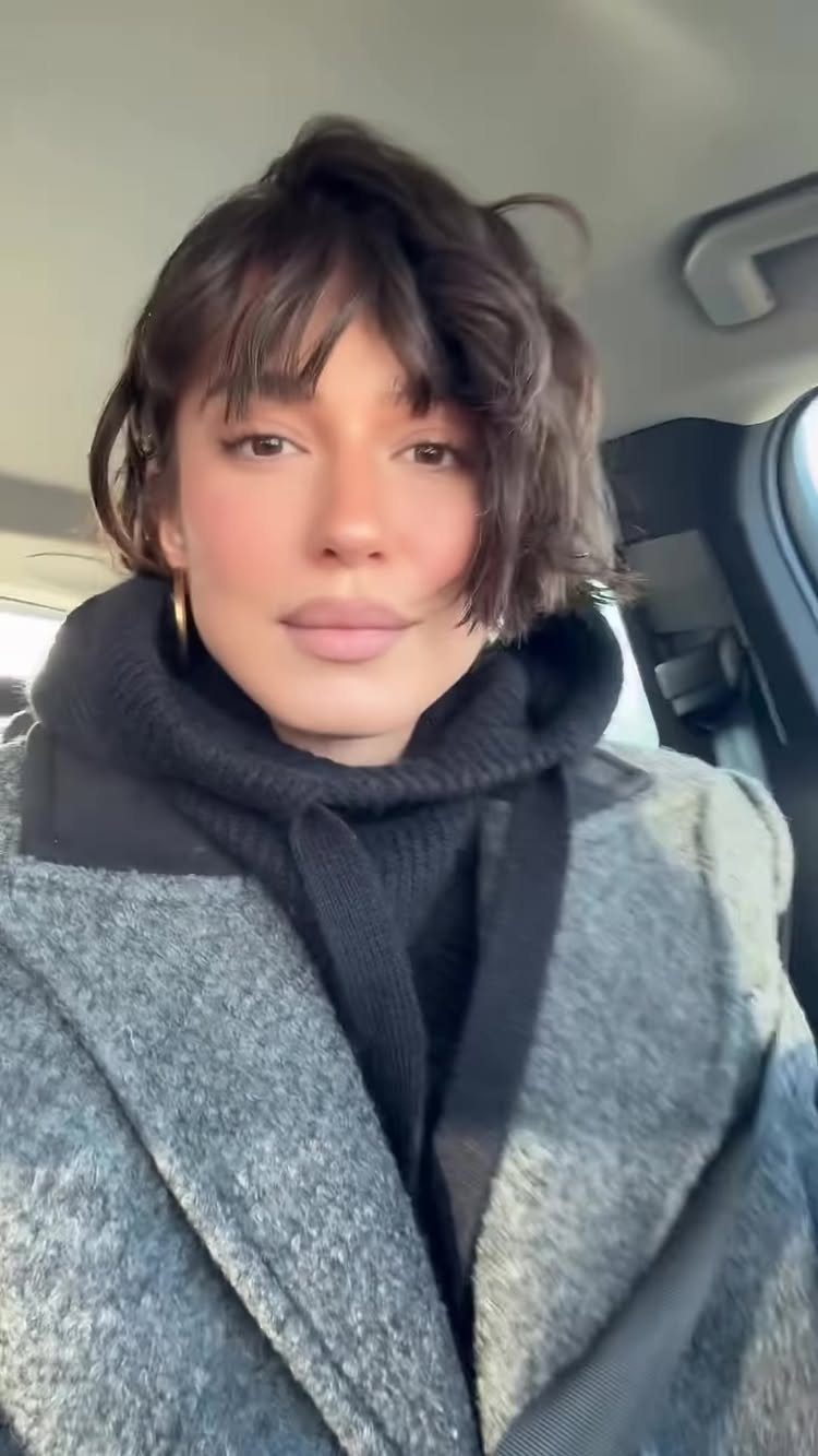Short tousled dark bob with wispy bangs framed by a gray coat and scarf