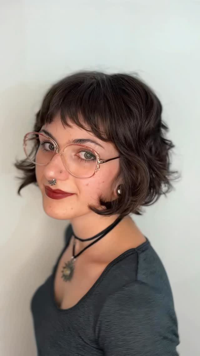 Rounded dark bob with wispy bangs, worn with glasses and a black choker