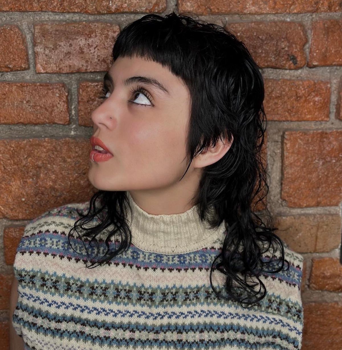 Person with a short blunt fringe and long wavy black mullet against a brick wall
