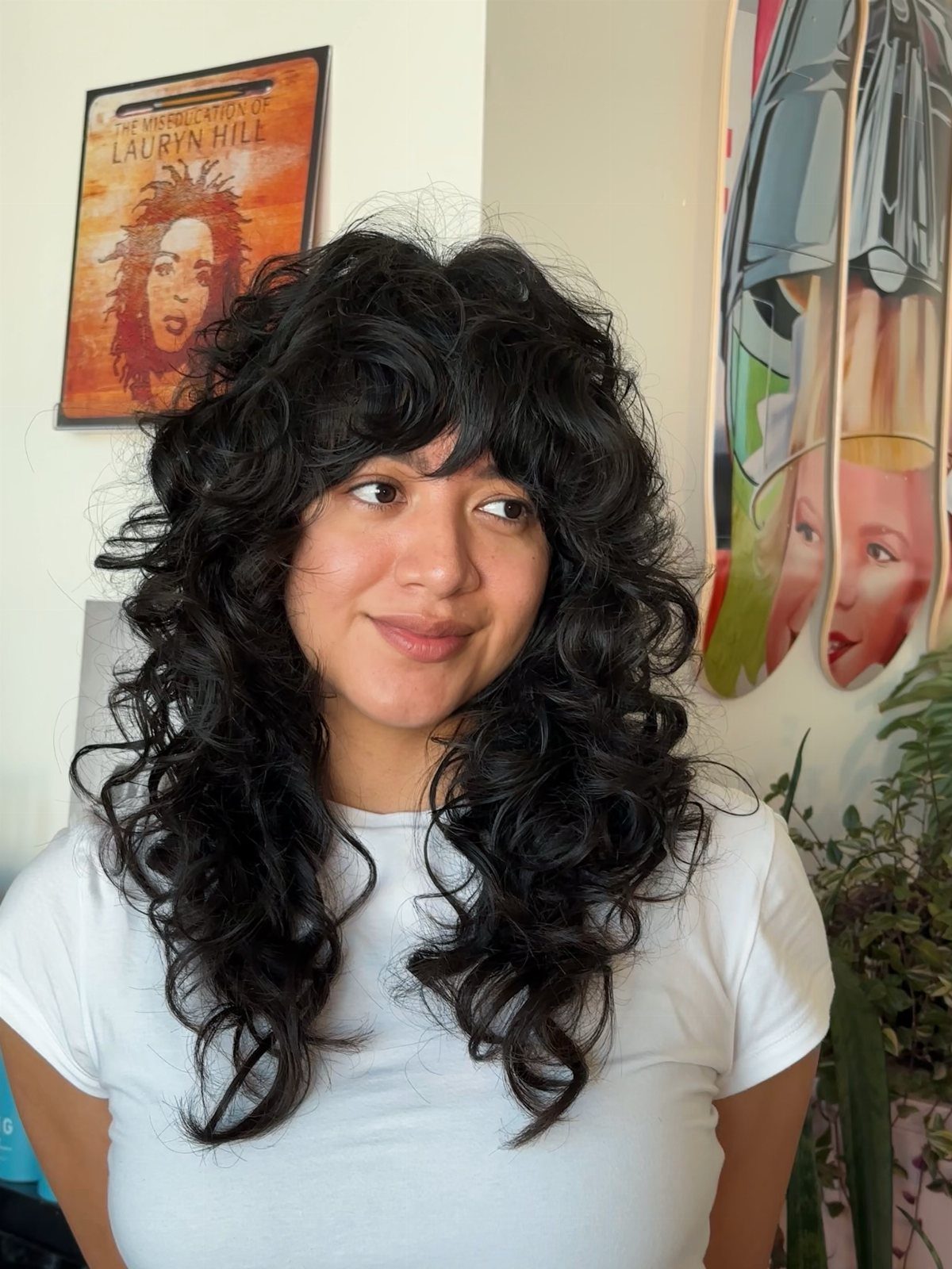 Long dark curls with tousled bangs frame a person wearing a white T-shirt