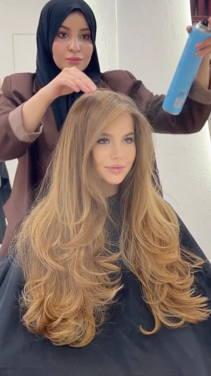 Long blonde layered hair falls in voluminous waves over a black salon cape
