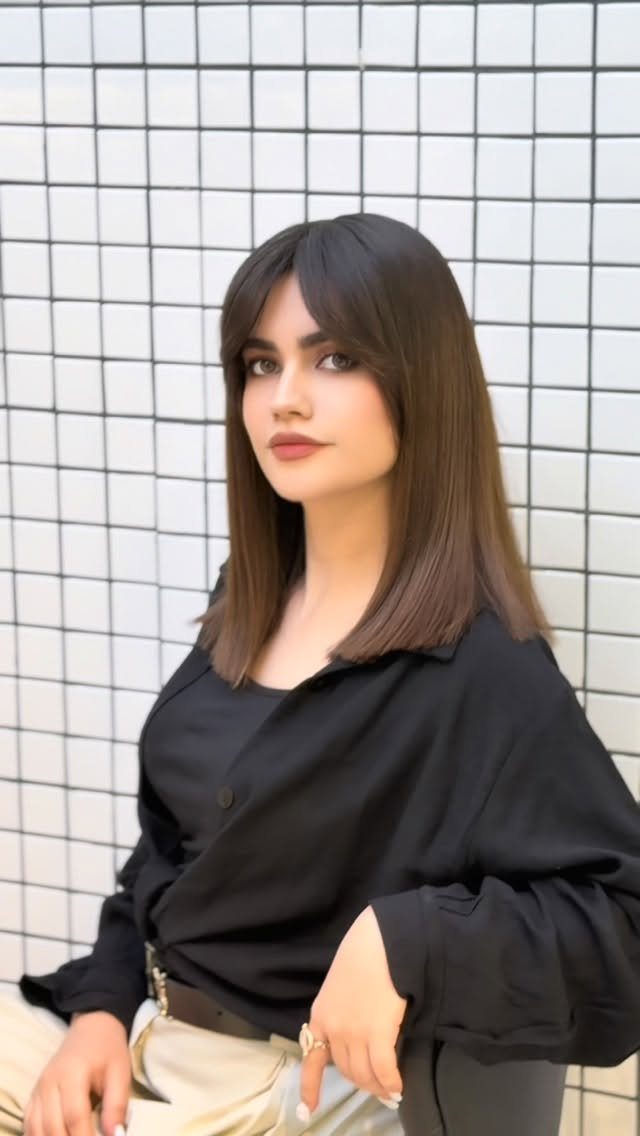 Woman with long straight dark hair and bangs posing against a tiled wall