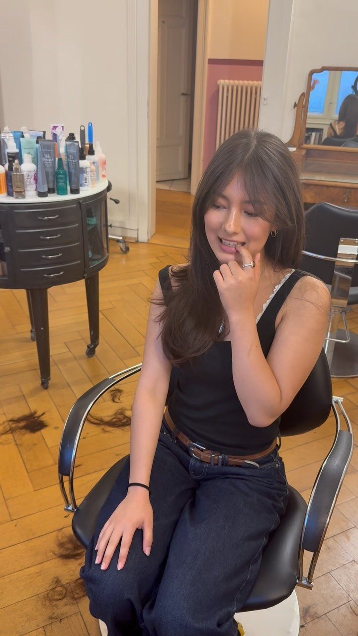 Woman with long dark hair and wispy face-framing bangs seated in a salon chair