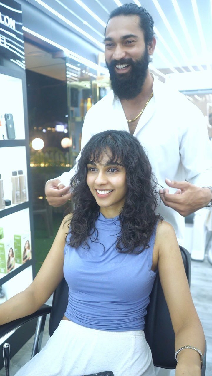 Woman with long dark curls and curly bangs seated in a salon chair