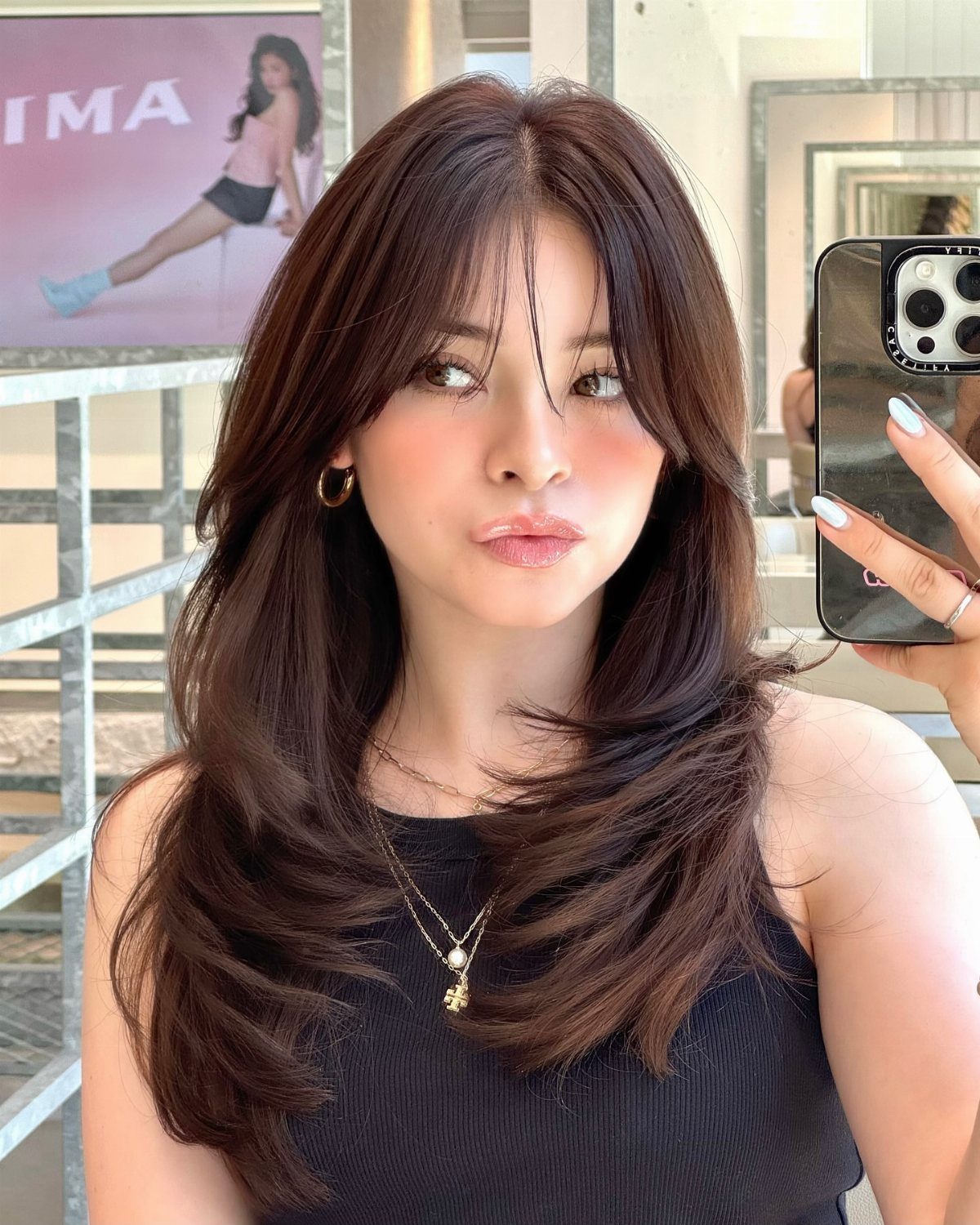 Woman with long layered brown hair and soft feathered bangs taking a mirror selfie