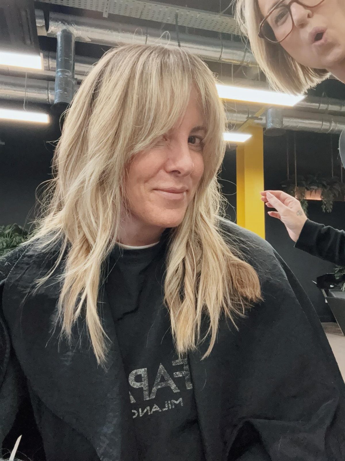 Woman with layered blonde hair and curved bangs seated in a salon