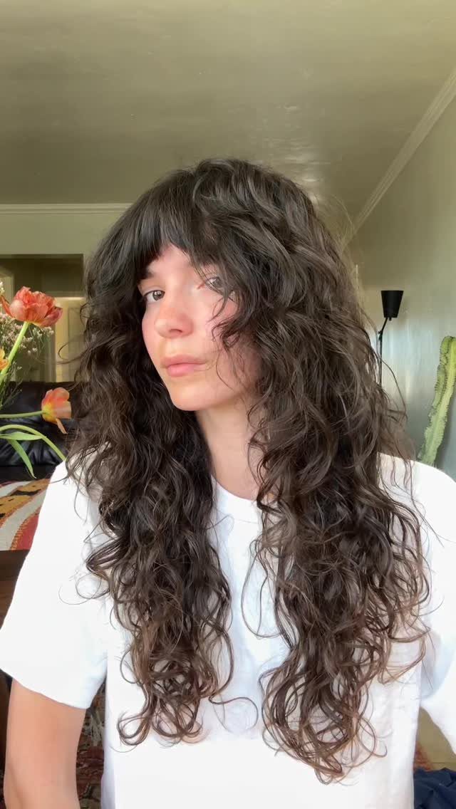 Person with long dark curls and curly bangs wearing a white T-shirt indoors