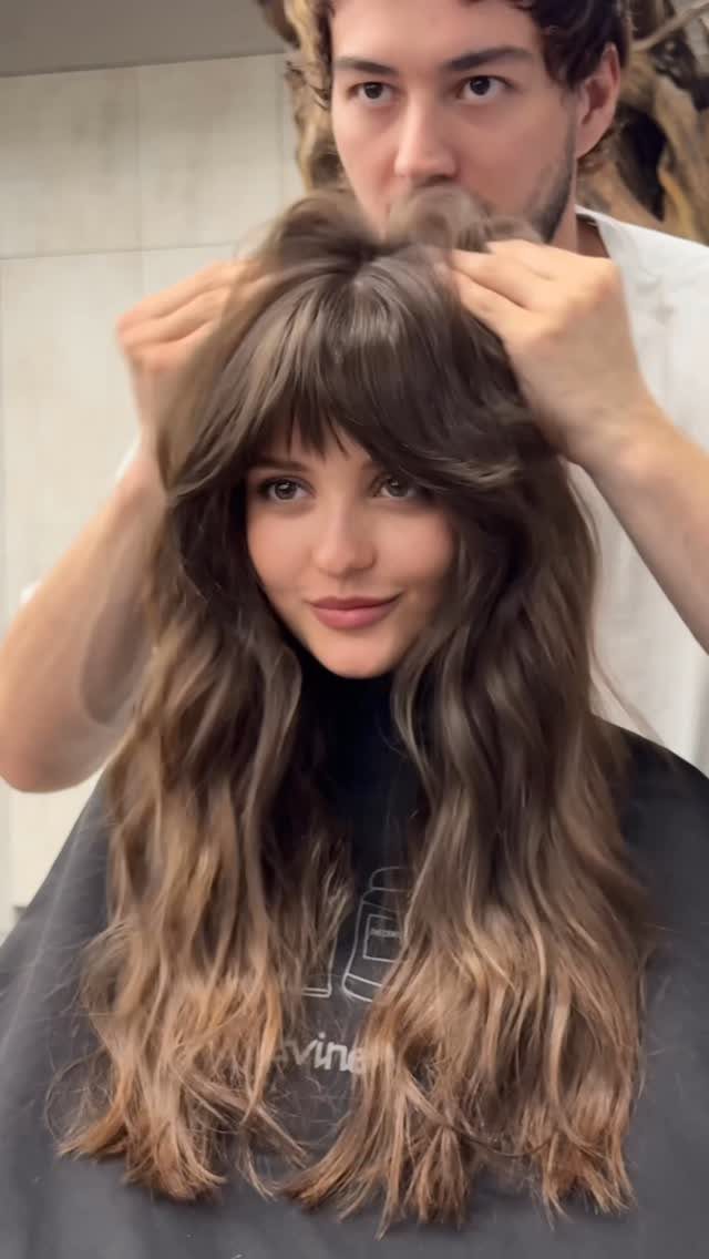 Woman with long wavy brown hair and wispy bangs as a stylist adjusts her hair