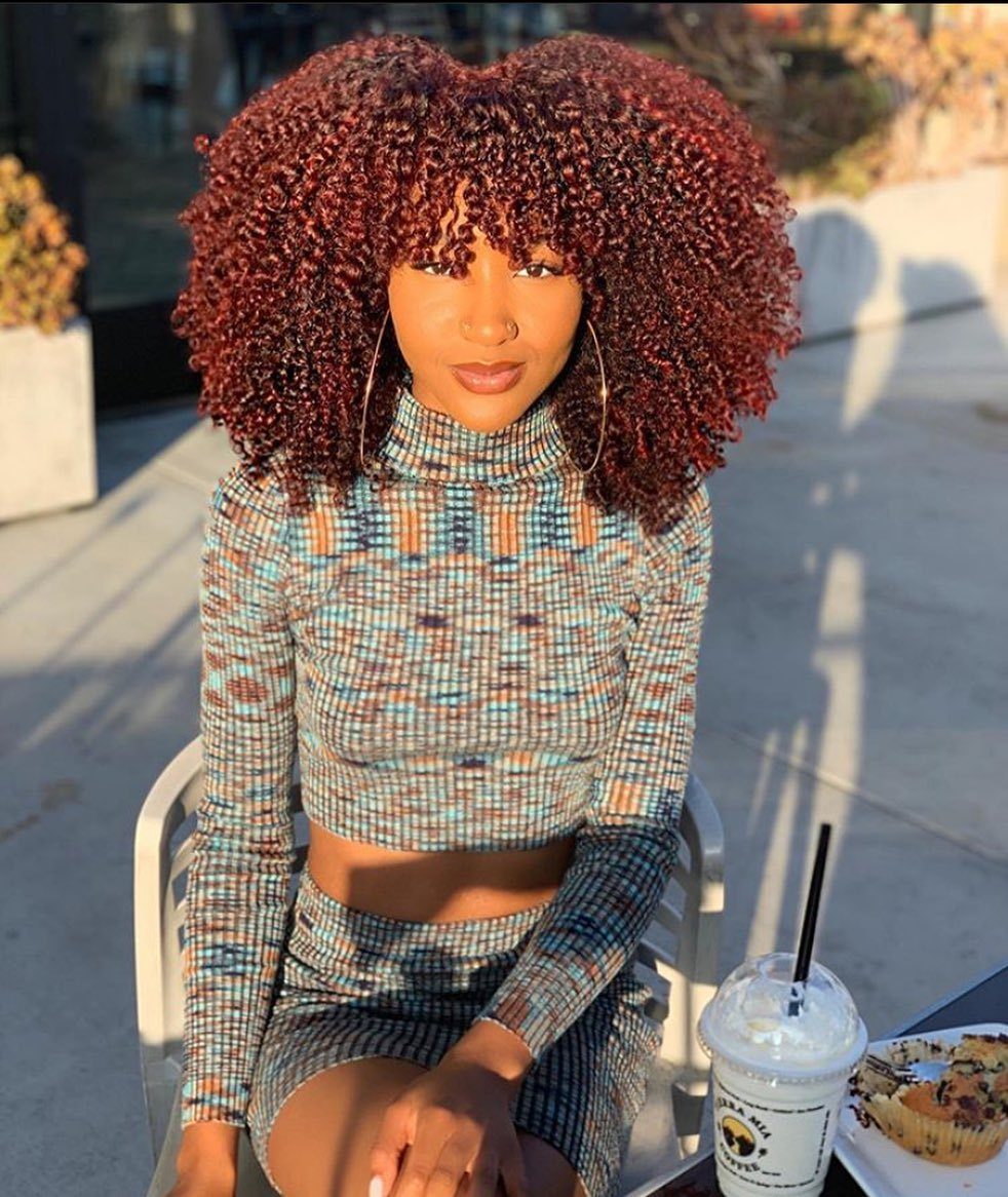 Woman with voluminous reddish-brown natural coils and bangs seated outdoors