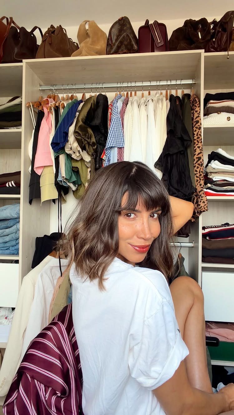 Woman with dark wavy shoulder-length hair and wispy bangs poses by a wardrobe