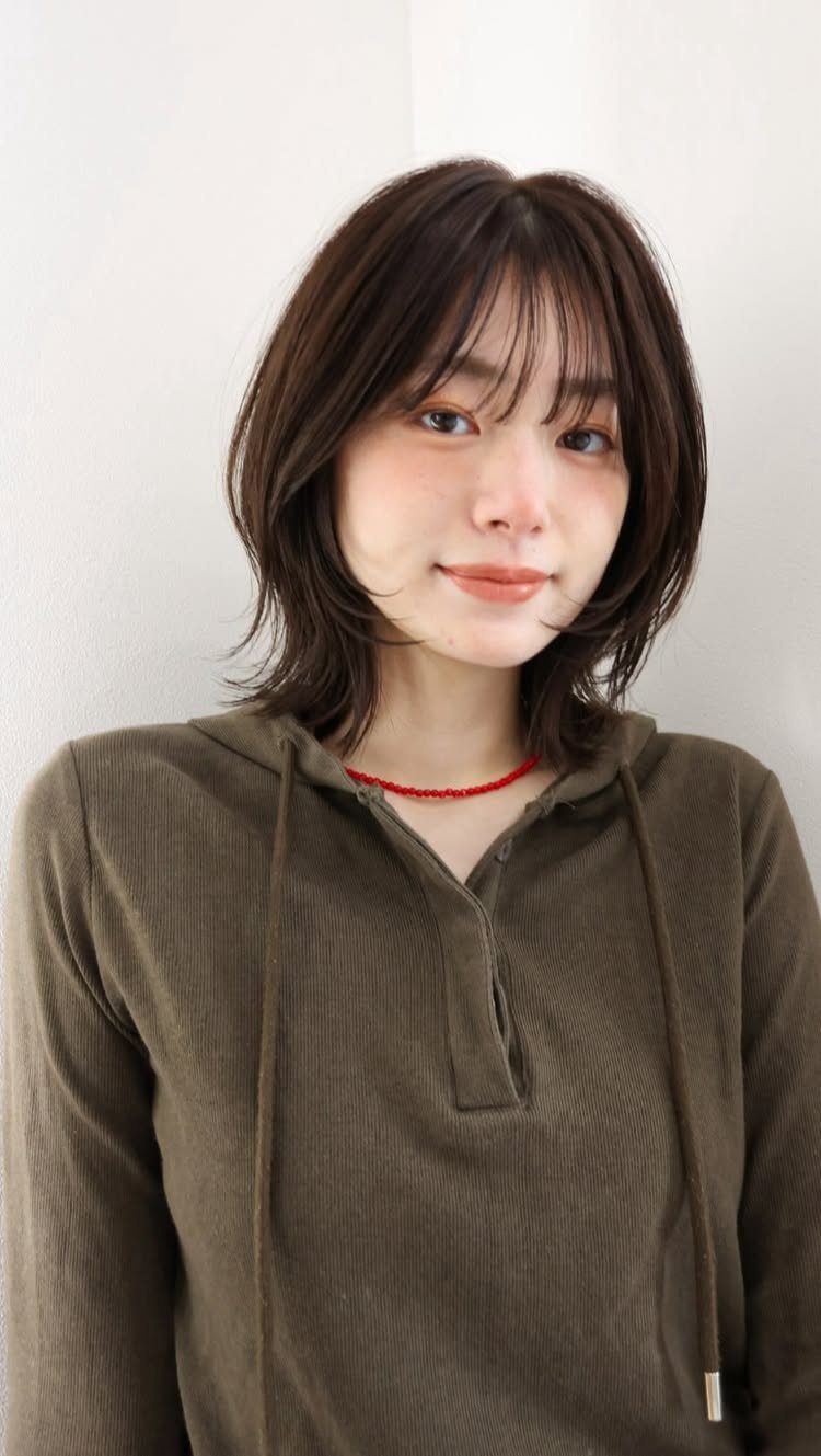 Woman showcasing a dark layered lob with wispy bangs against a plain indoor wall