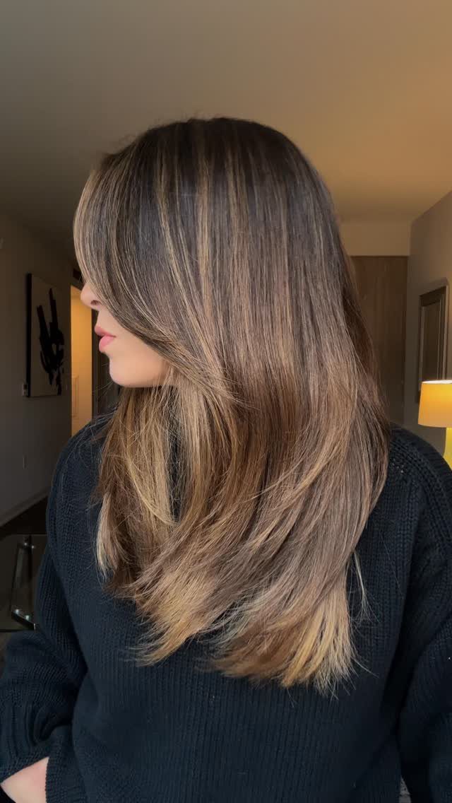 Long brown hair with lighter ends falls in soft layers over a black sweater