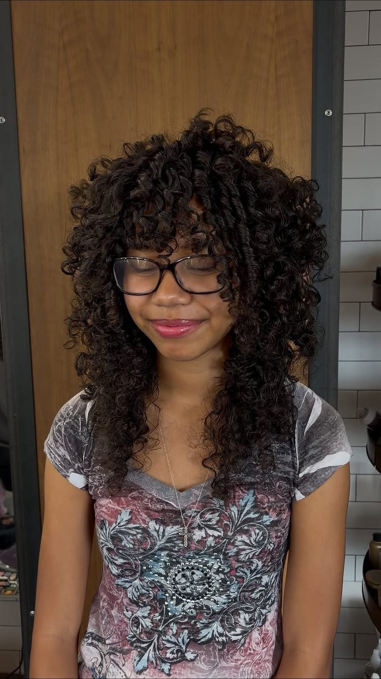 Shoulder-length dark curls with layered volume and bangs framing a person’s face