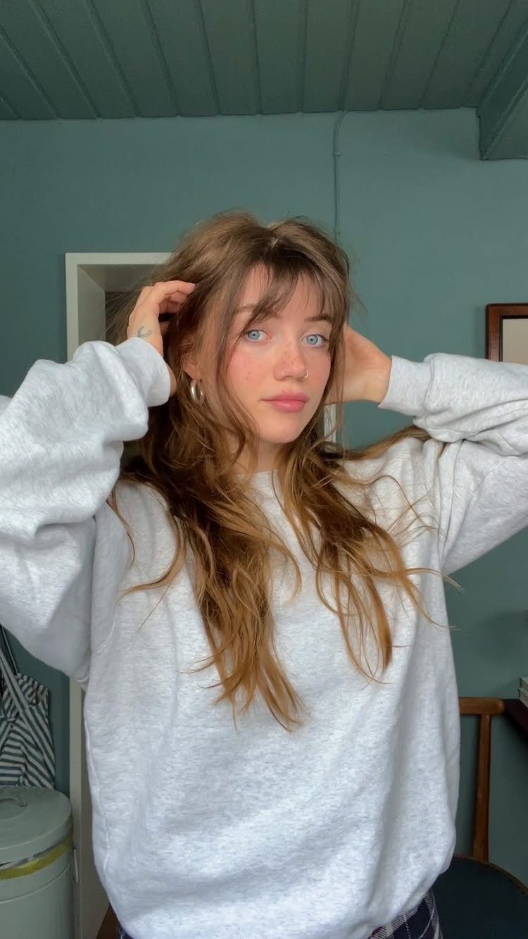 Woman with long, softly wavy brown hair and wispy bangs in a gray sweatshirt