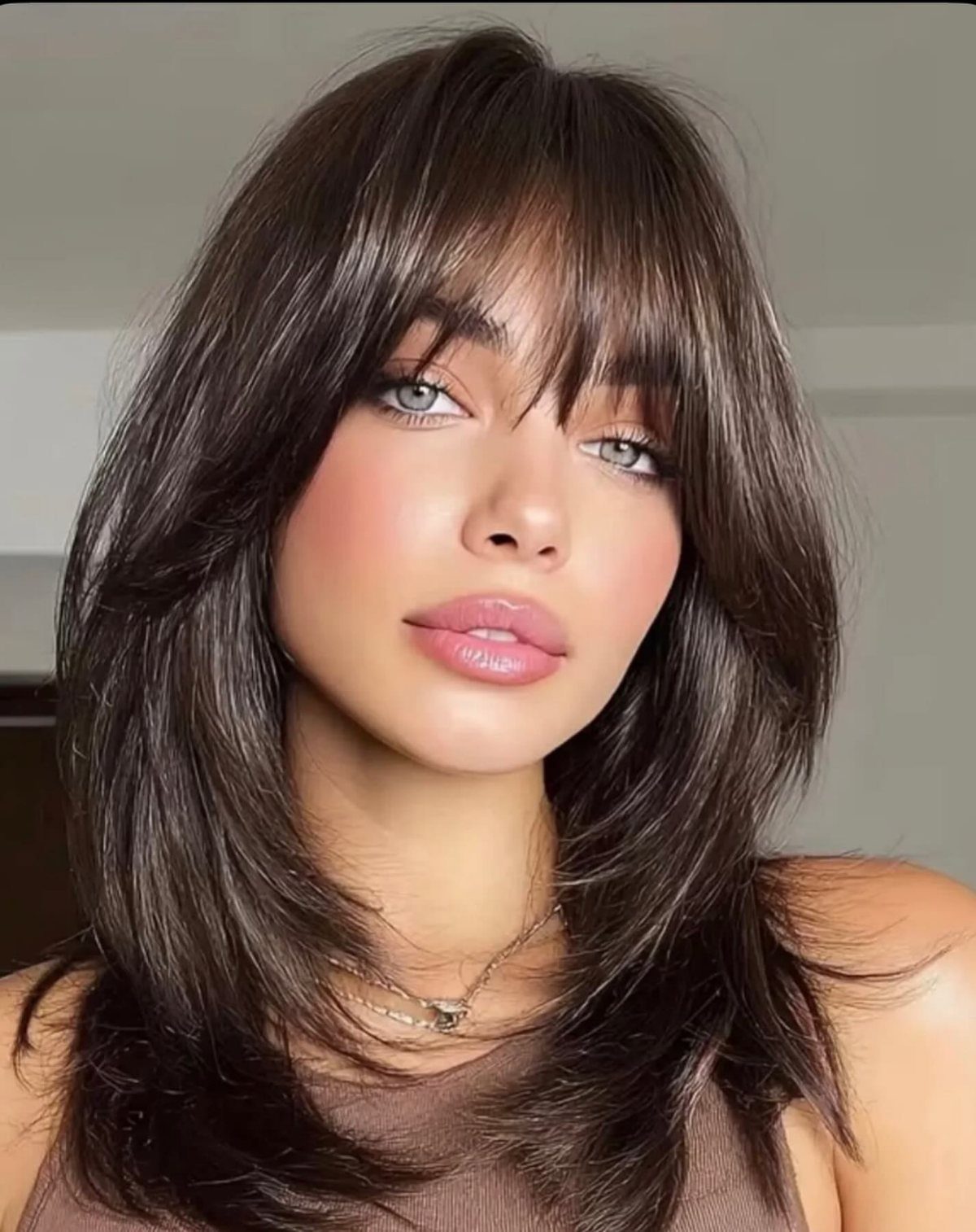 Woman with glossy dark layered hair, feathered ends, and wispy bangs