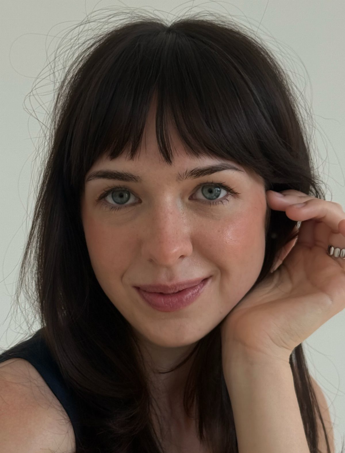 Woman with long dark layers and short uneven bangs posing against a pale wall