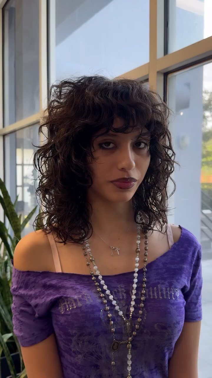 Person with a dark curly shag haircut, layered shoulder-length curls, and bangs