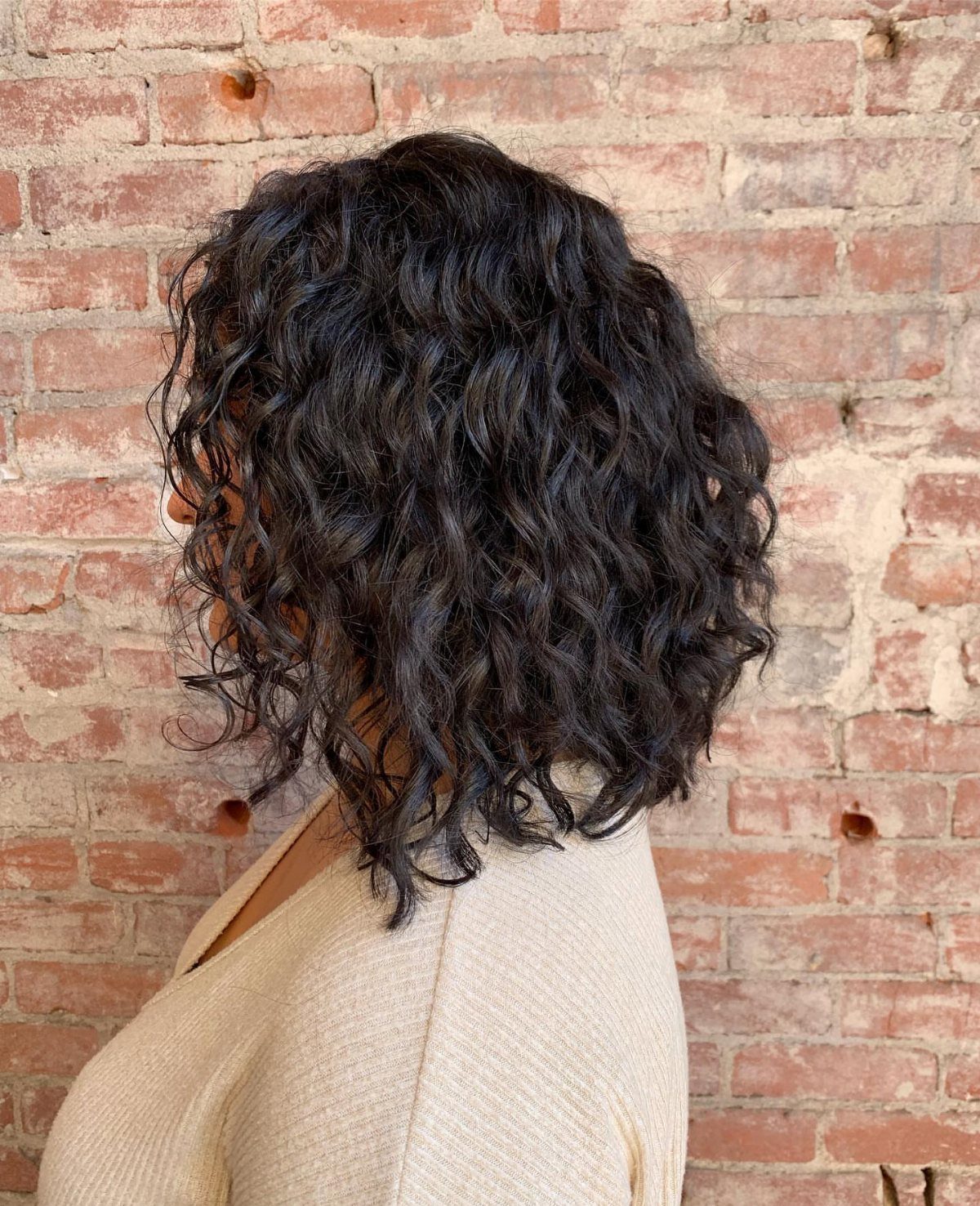 Shoulder-length dark curls forming a rounded lob against a weathered brick wall