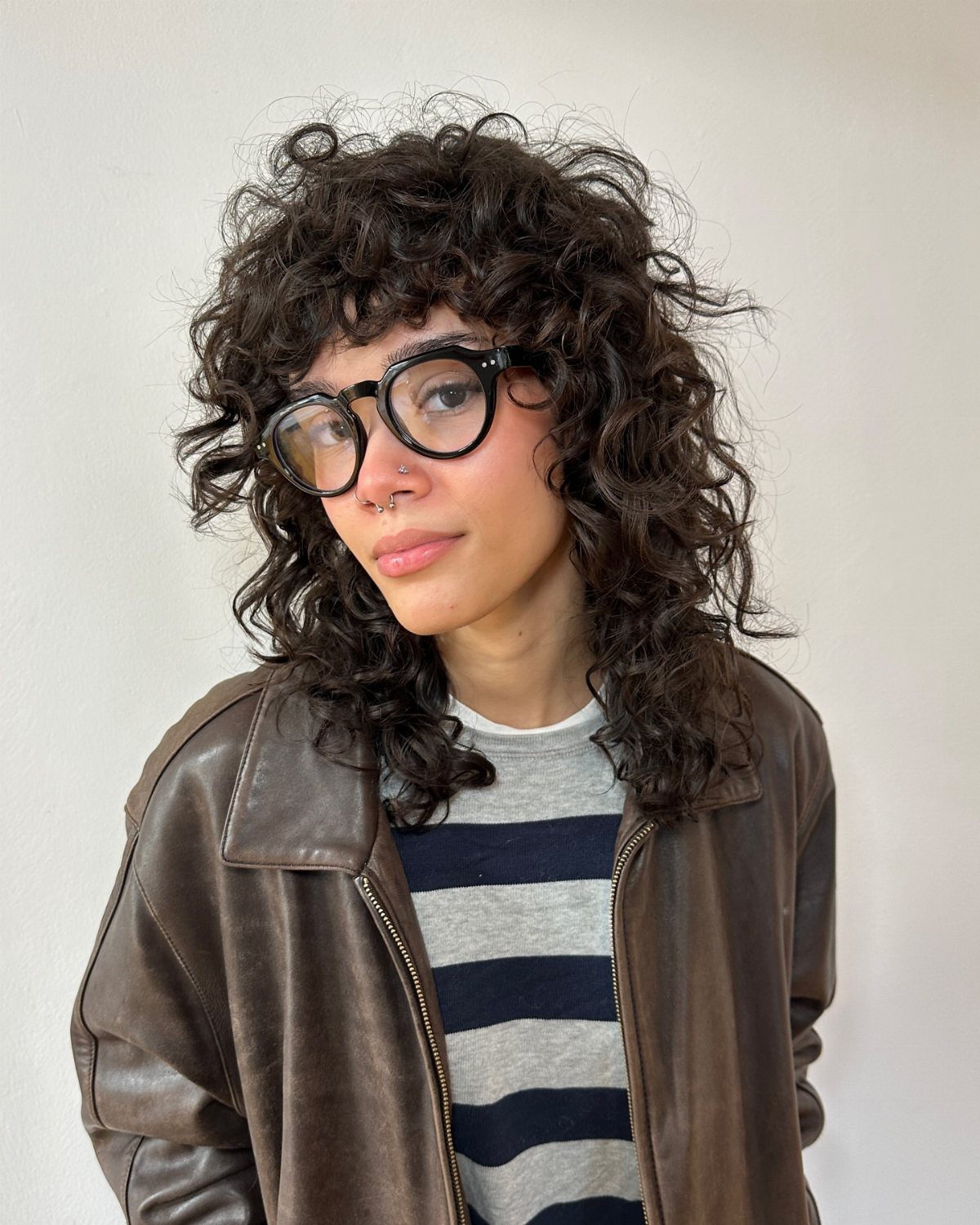 Person with voluminous dark curls, curly fringe, black glasses, and brown leather jacket