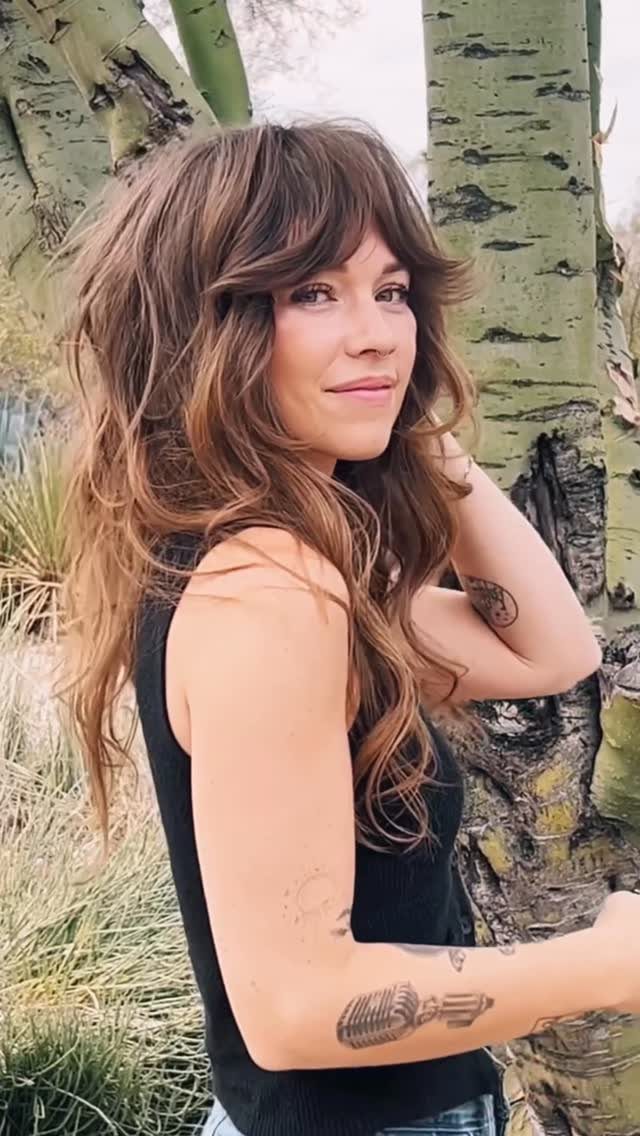 Woman with long shag layers and wispy bangs posing beside birch trees