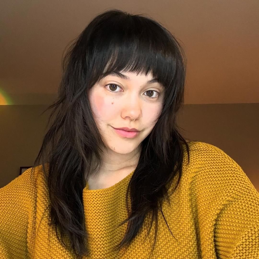 Person wearing a mustard sweater with long dark shaggy hair and full bangs