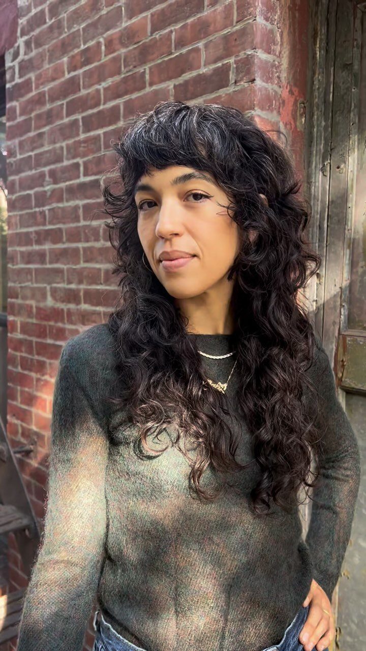 Person with long dark wavy shag and bangs standing against a brick wall
