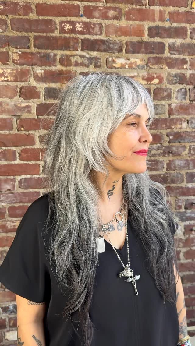 Woman with long silver-gray shag haircut and bangs against a brick wall
