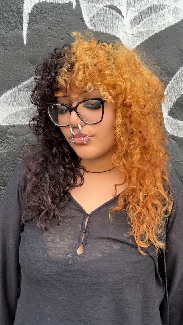 Person with long, thick split-color curls wearing glasses against a gray wall