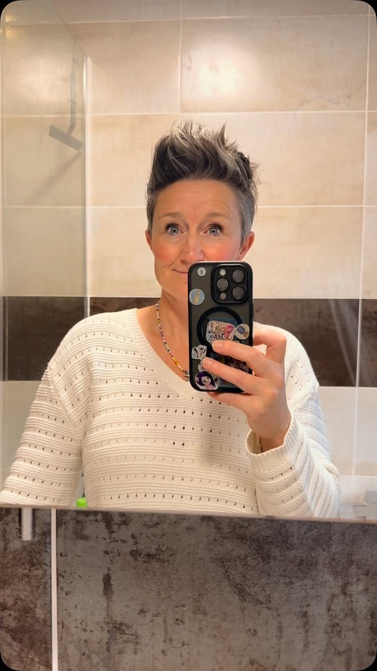 Person with short dark hair styled upward in a quiff, taking a bathroom mirror selfie
