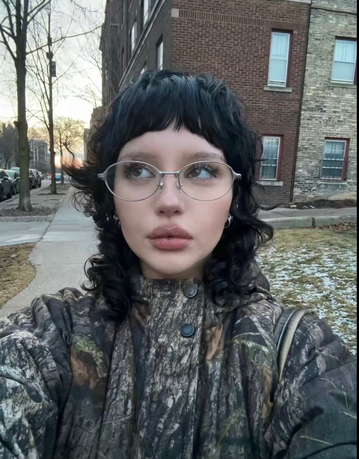 Person with short dark layered hair, choppy bangs, round glasses, and camouflage coat