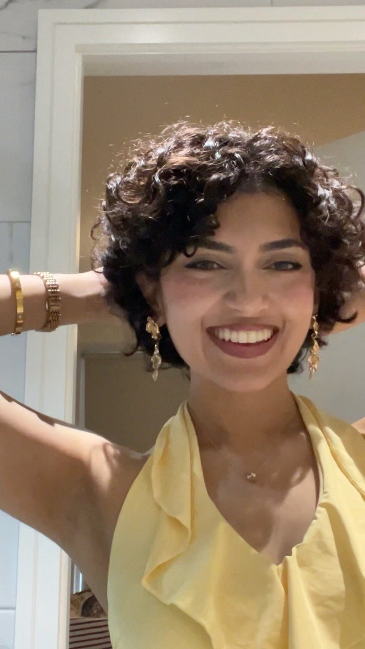 Woman smiling with short dark curls and an upward-flipped side piece