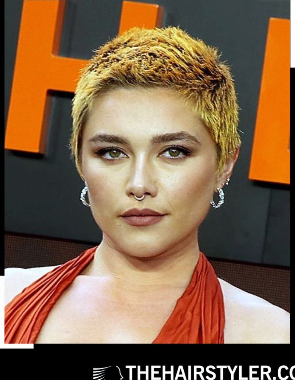 Woman with a short textured blonde pixie cut, silver earrings, and an orange draped top.